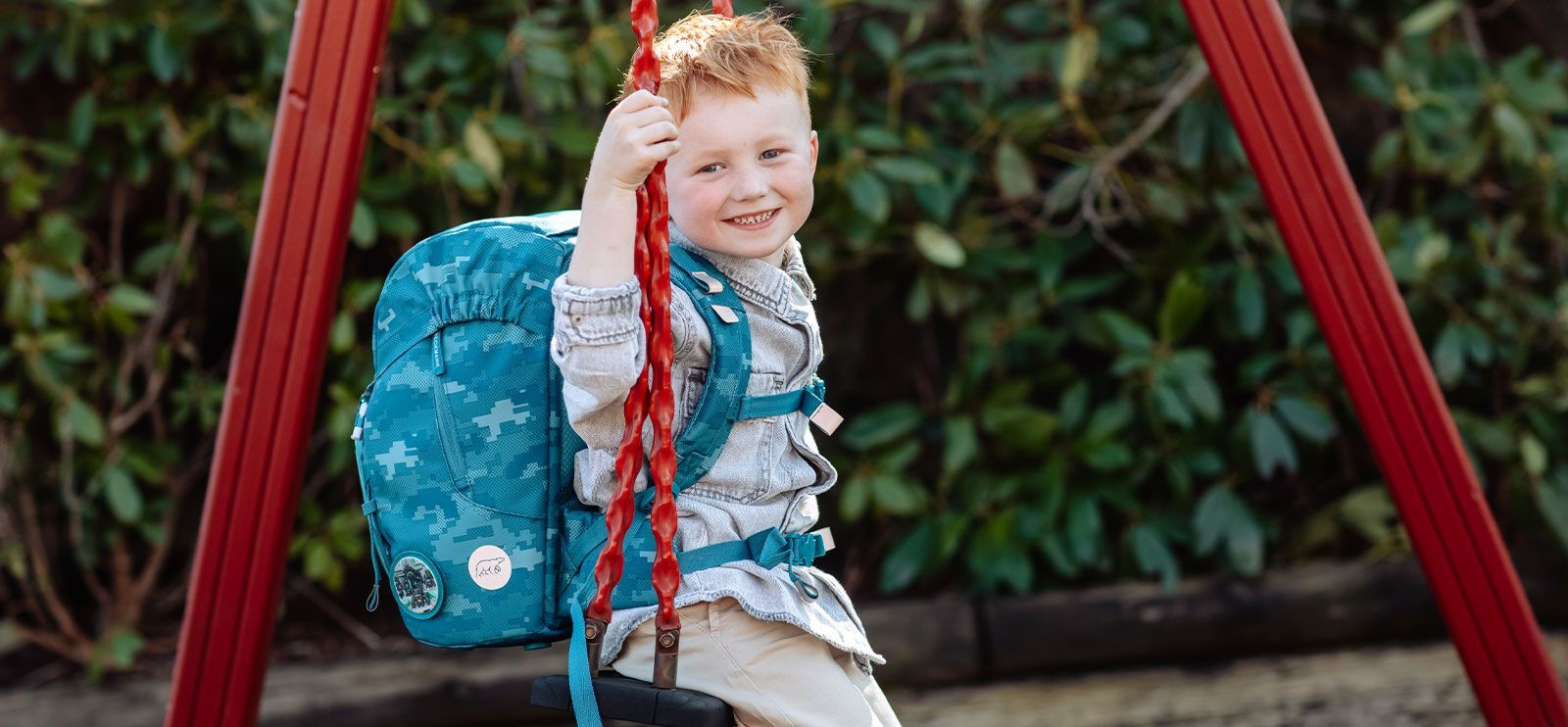 Liten gutt i blomstereng, han lukter på en blomst. Han har på seg rød genser og blå ryggsekk.
