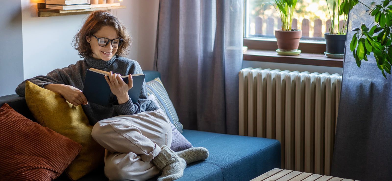 Dame med brunt hår og briller sitter i en sofa og koser seg med en god bok.
