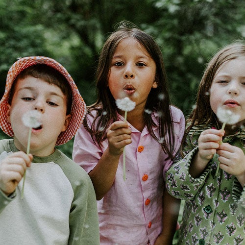 Fæbrik kids Fæbrik kids: Tre barn i hjemmesydde klær ser i kameraet og blåser på en løvetann.