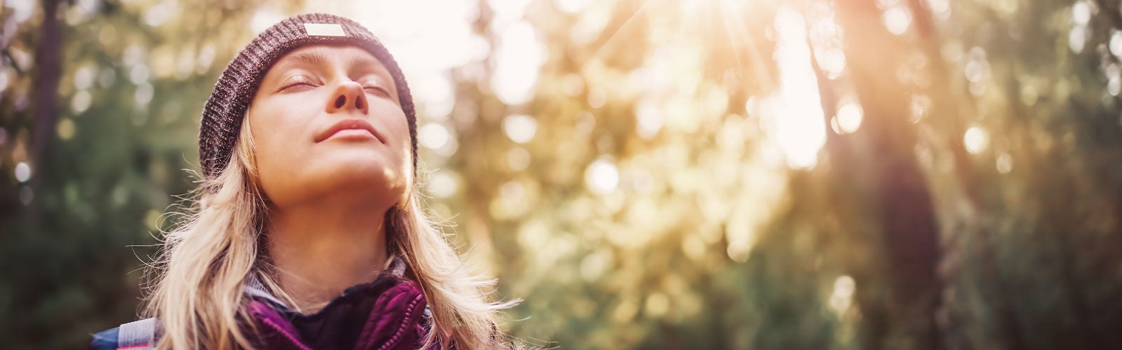 Blond kvinne med lue står i skogen. Hun lukker øynene og har ansiktet vendt mot himmelen.